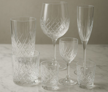 Collection of luxury drinking glasses in various sizes, crystal clear, placed on marble surface with soft lighting, showcasing intricate patterns and elegant shapes
