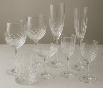 Complete crystal glass set arranged beautifully on a white linen tablecloth, featuring various sizes of wine glasses, champagne flutes, and cocktail glasses with sparkling reflections